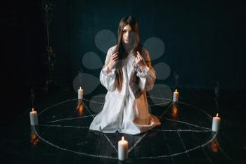 Young woman in white shirt sitting in the center of pentagram circle with candles and reads a spell, black wooden floor, smoke all around. Dark magic ritual, occultism