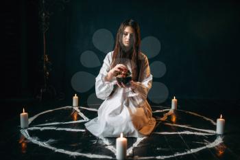 Young woman in white shirt sitting in the center of pentagram circle with candles and produces a ritual of black magic, black wooden floor. Occultism and exorcism