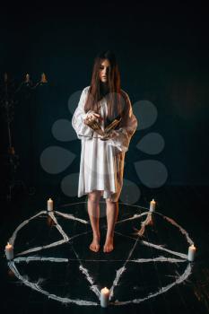 Young woman in white shirt standing in the center of pentagram circle with candles and reads a spell, black wooden floor. Dark magic ritual, occult and exorcism