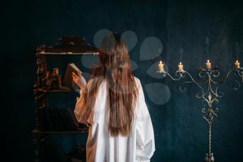 Female person in white shirt puts old spellbook on shelf, back view, candles on background. Dark magic, occultism and exorcism, witchcraft