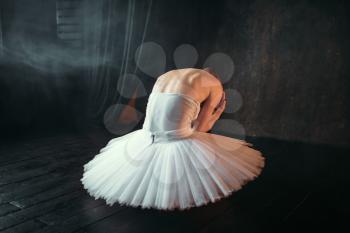 Classical ballet dancer in white dress sitting on theatrical stage, back view. Ballerina training in class with black background