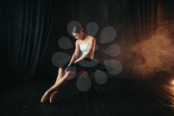 Ballet dancer sitting on black banquette on the stage in theatre. Graceful ballerina training in class