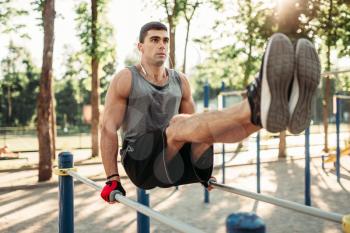 Athletic man in headphones doing exercise on press using parallel bars, outdoor fitness workout. Strong sportsman on sport training in park