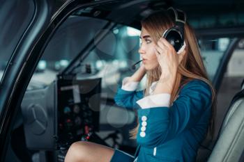 Female pilot in headphones poses in helicopter cabin, hangar interior on background, view from cabin. Air hostess in uniform sits in copter. Private airline