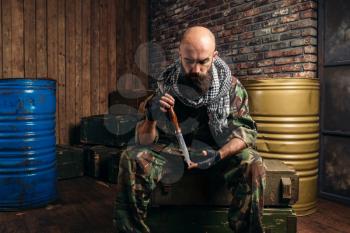 Terrorist in uniform with knife in hands sitting on boxes of ammunition. Terrorism and terror, soldier in camouflage in weapon arsenal, barrels of fuel or chemicals on background