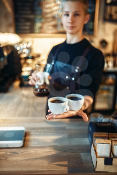 Young male barista in black apron holds in hands two cups of fresh coffee, cafe counter on background. Professional espresso preparation by barman in cafeteria, bartender occupation