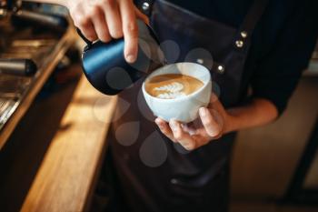 Barista hand pours cream into the cup of coffee, wooden table on background. Professional cappuccino preparation by bartender