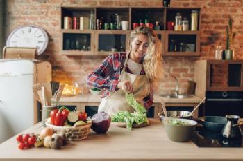 Housewife in apron prepares salad, kitchen interior on background. Female cook making healthy vegetarian food, vegetables preparation