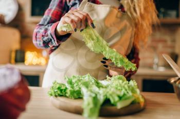 Housewife in apron prepares salad, kitchen interior on background. Female cook making healthy vegetarian food, vegetables preparation
