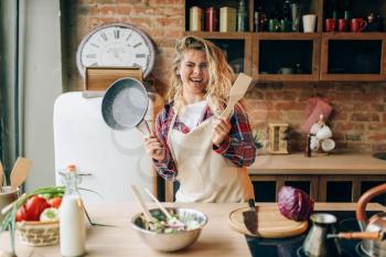 Smiling housewife in an apron holding frying pan and wooden spatula in hands, kitchen interior on background. Female cook making healthy vegetarian food, salad cooking