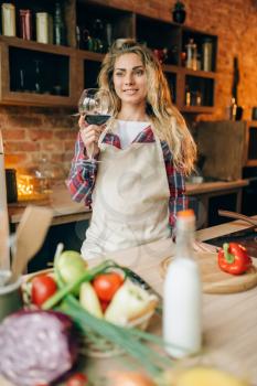 Happy housewife in an apron holding glass of red wine in hands, kitchen interior on background. Female cook before vegetable salad preparation