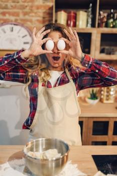 Playful housewife in an apron covered her eyes with eggs, kitchen interior on background. Female cook prepares fresh homemade cake. Domestic pie preparation