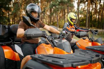 Two quad bike riders in helmets and equipment, side view, closeup, summer forest on background. Male quadbike drivers, atv riding, extreme sport