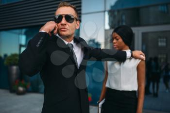 Serious bodyguard in suit and sunglasses requests support on earpiece for female client protection. Security guard is a risky profession, professional guarding