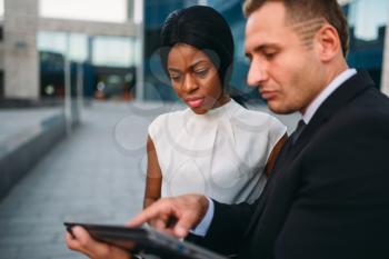 Business woman and businessman looks on laptop screen, outdoors meeting of partners, modern office building on background, partnership negotiations during the lunch break