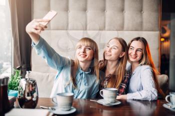 Three happy girlfriends makes selfie on phone camera in cafe. Female friends sitting in restaurant and having fun, gossipers meeting