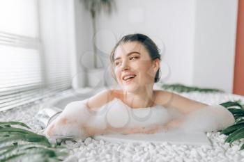 Young smiling woman in bathtube with foam. Luxury bathroom with window and palm branch decor