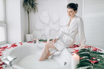 Elegant woman in white bathrobe sitting on the edge of the bath decorated with rose petals. Luxury bathroom interior with window on background