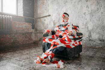 Autistic wraps in crime-scene tape and sitting in the middle of the room. Mad male person in abandoned house, strange guy