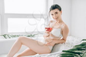 Female person sitting in the bath with glass of red wine. Bathroom interior with window on background