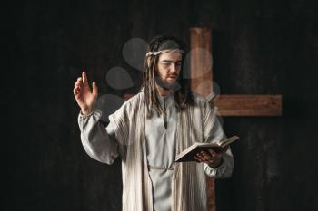 Holy Jesus Christ praying with biblical in hands, cross on black background. Prayer against crucifixion, christianity symbol