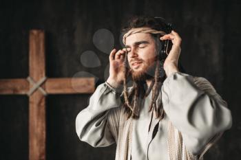 Man in the image of Jesus Christ listening to music with headphones, crucifixion cross on black background. Believers and modern technologies concept