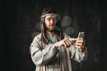 Man in the image of Jesus Christ holds mobile phone, dark background. Gadgets from the evil one