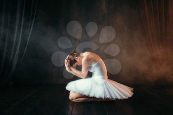 Ballerina in white dress sitting on theatrical stage, side view. Classical ballet dancer training in class with black background