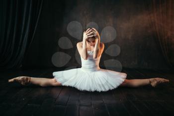 Body flexibility of ballet performer, stretching exercise. Ballerina in white dress sits on a twine on black wooden floor