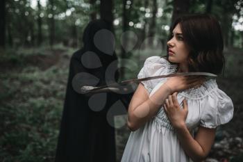 Death in a black hoodie swinging a scythe on young female victim, forest on background. Photo in horror style, mystery ritual