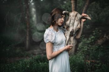Female victim in white dress with skull of the animal instead of the head, forest on background. Photo in horror style, exorcism