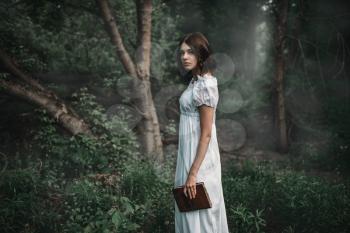 Female victim in white dress holds book in hand, forest on background. Photo in horror style, mystery