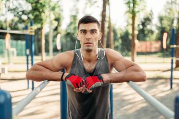 Athletic man in headphones doing exercise on parallel bars, front view, outdoor fitness workout. Strong sportsman on sport training in park