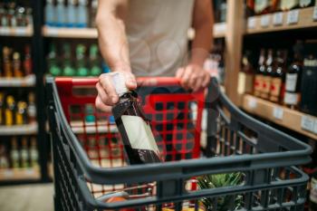 Male person put bottle of wine in a cart, alcohol section in market. Shelves with drinks on background