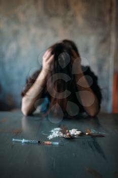 Female junkie sitting at the table with drugs and syringe, grunge room interior on background. Drug addiction concept, addicted people