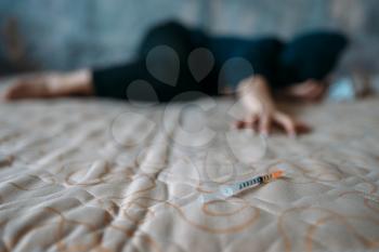 Female druggy with syringe in hand lies in bed, bottle of alcohol and spoon for dose preparing on background. Drug addiction concept, addicted people