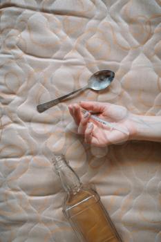 Female junkie hand with syringe against bottle of alcohol and spoon for dose preparing on background. Drug addiction concept, addicted people