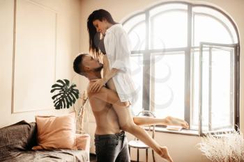 Husband holding wife in his arms against window with sunlight, erotica in bedroom. Erotic scene, sexual relationship of love couple