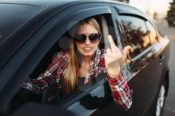 Female car driver in sunglasses shows an obscene gesture. Impudent woman in vehicle, aggressive automobile driving concept