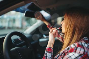 Young woman driver paints her lips with lipstick in a car. Female person preens in vehicle, driving automobile concept