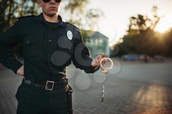 Serious police officer in uniform and sunglasses holds hand handcuffs in hands, front view. Cop at the work. Law protection concept, safety control job