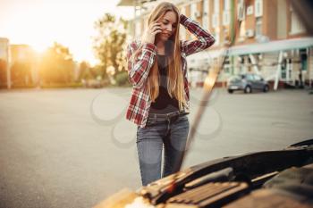 Car breakdown concept, sad woman against open bonnet on the roadside. Female driver looks at the broken vehicle engine