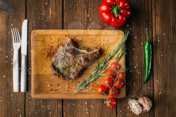 Juicy grilled steak on the bone, top view, nobody. Knife and fork on wooden table on background, meat dish