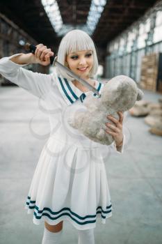 Anime style blonde girl with glass face cuts off the head of a teddy bear. Cosplay fashion, asian culture, doll with blade, cute woman with makeup in the factory shop