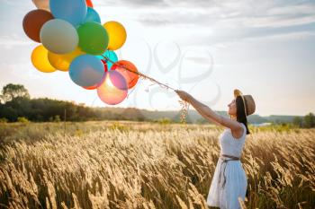 Pretty girl releases a bunch of balloons into the sky in wheat field, back view. Cute woman on summer meadow at sunny day