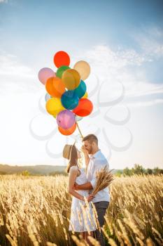 Happy love couple with balloons hugs in wheat field. Pretty wife and husband walking on summer meadow