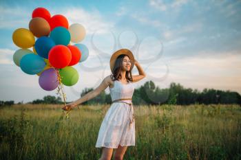 Smiling woman with colorful balloons walking in green field. Pretty girl on summer meadow at sunny day