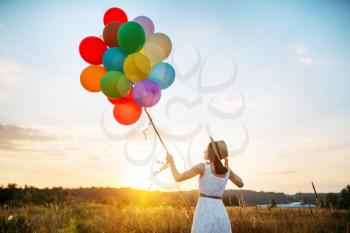 Girl with colorful air balloons walking in wheat field. Pretty woman on summer meadow at sunny day