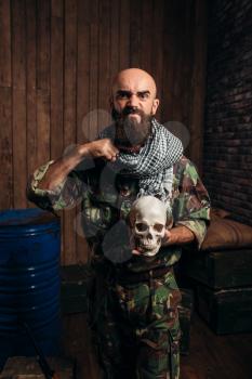 Terrorist in uniform holds human skull in hands, male mojahed. Terrorism and terror horror, bearded soldier in khaki camouflage, barrels of fuel or chemicals on background
