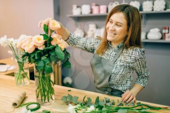 Female florist in apron puts fresh roses in a vase in flower shop, floral business concept